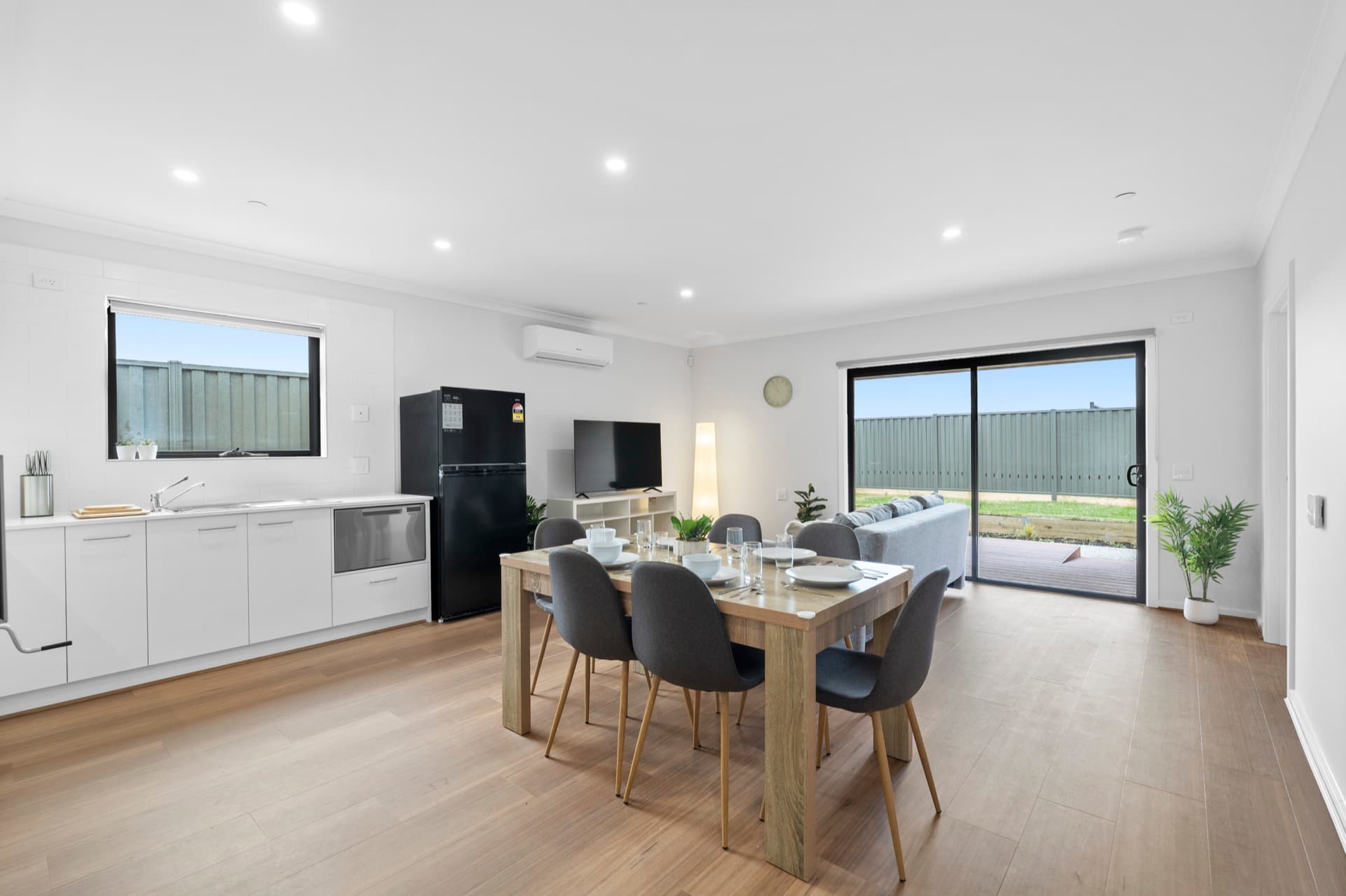 Open plan dining and kitchen area with six-seater timber table, white cabinetry, and black fridge