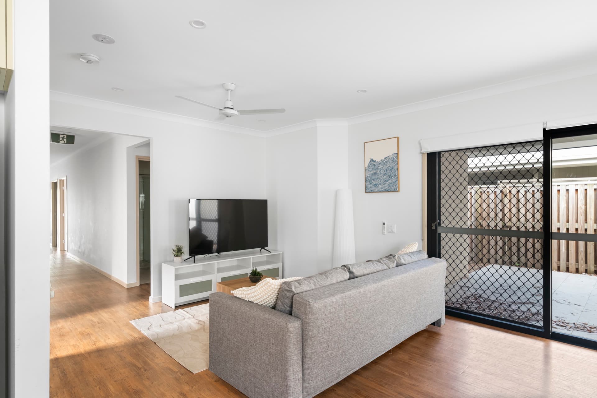 Living room with grey sofa, ceiling fan, TV on white entertainment unit, and hallway leading to bedrooms